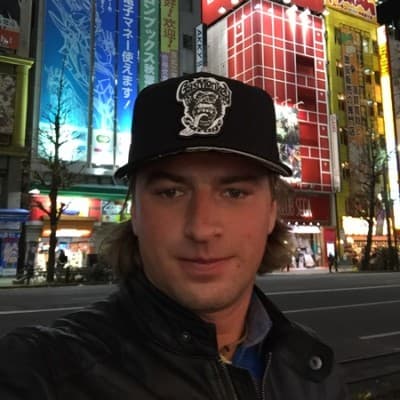 Man in a Gas Monkey cap and leather jacket before neon city signs at night.